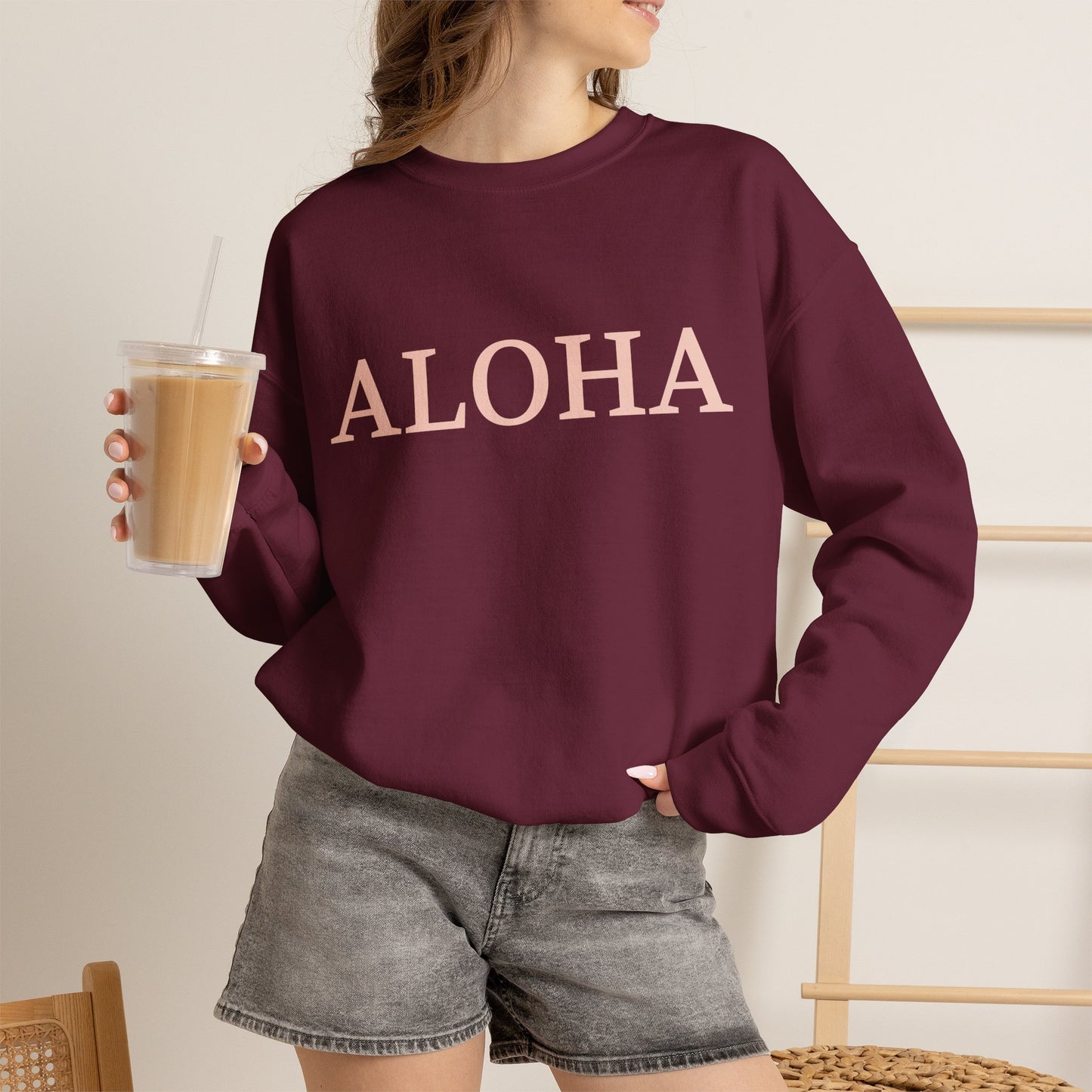 A woman wearing an oversized crewneck sweatshirt in deep burgundy with the word 'ALOHA' printed across the chest in a light pink color. She is holding a clear plastic cup of iced coffee and wearing gray denim shorts, set against a warm, neutral indoor background.