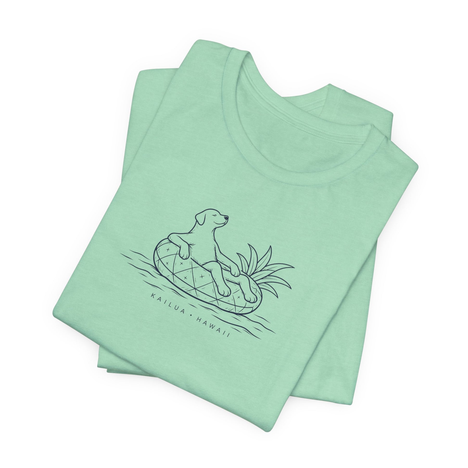 A folded mint green t-shirt featuring a black line art graphic of a dog sitting on a pineapple in water, with the text 'KAILUA - HAWAII' printed below.