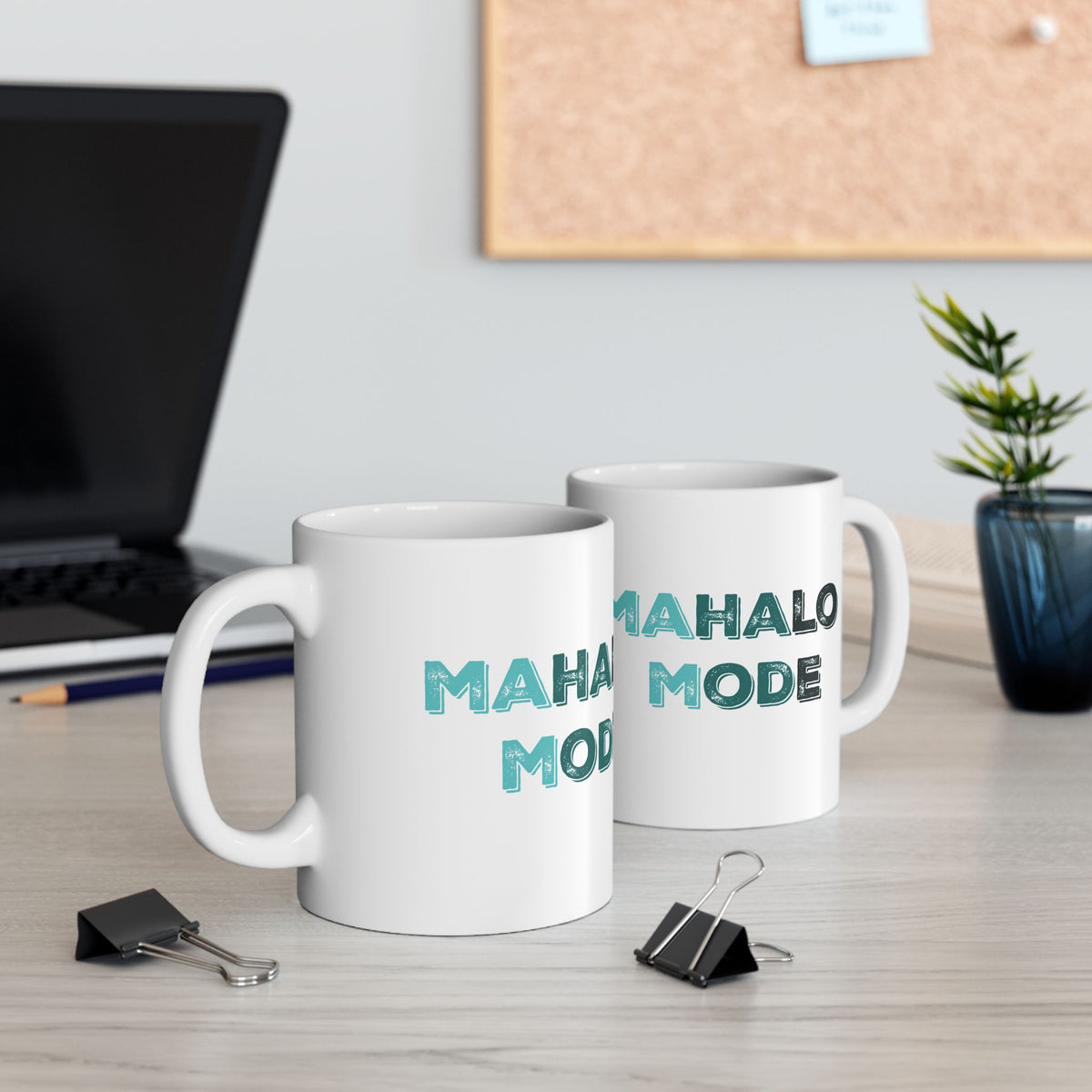 Two white ceramic mugs printed with the phrase 'MAHALO MODE' in teal lettering, displayed on a light wood desk in an office setting with a laptop, binder clips, and a plant in the background.