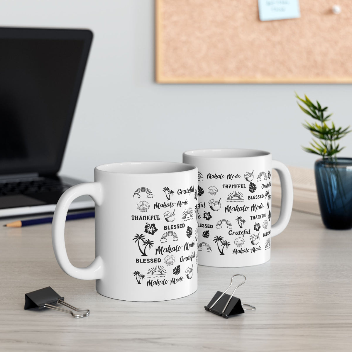 Two white ceramic mugs with black tropical-themed text and illustrations, displayed on a light wood desk in an office setting with a laptop, binder clips, and a plant in the background.