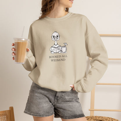 Woman wearing Sand sweatshirt with graphic print of a cartoon nene goose sitting reading a book with a cocktail next to it with text 'BOOKED ALL WEEKEND' holding an iced coffee