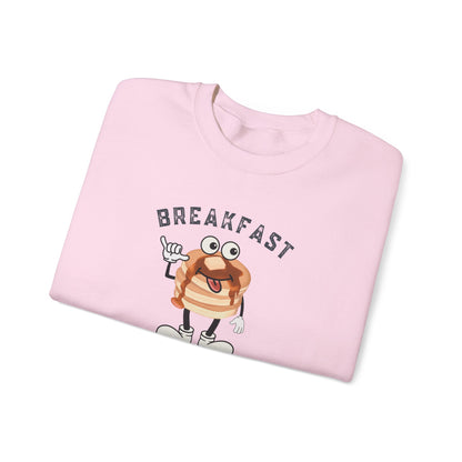 A folded light pink crewneck sweatshirt featuring a cartoon graphic of a smiling striped pancake with arms and legs, under the word 'BREAKFAST' in arched dark letters, shown on a plain white background.