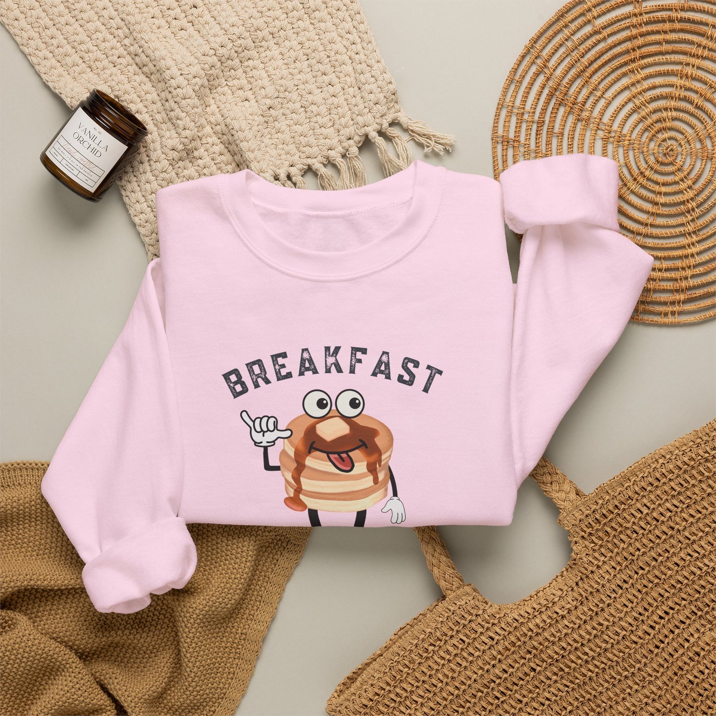 A light pink crewneck sweatshirt laid flat on a neutral surface, featuring a cartoon pancake graphic with the word 'BREAKFAST' above it. The sweatshirt is styled with a beige knit blanket, a small candle, and woven accessories.