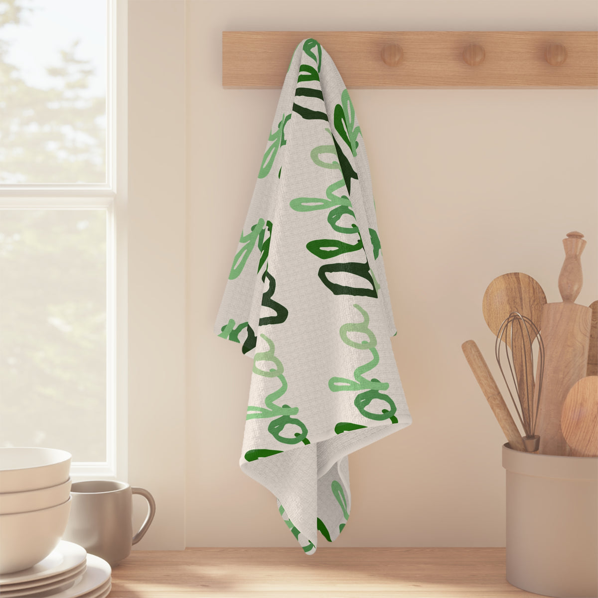 Green Aloha Hawaiʻi kitchen towel with repeating script design in light and dark green on white hanging off wooden peg in kitchen