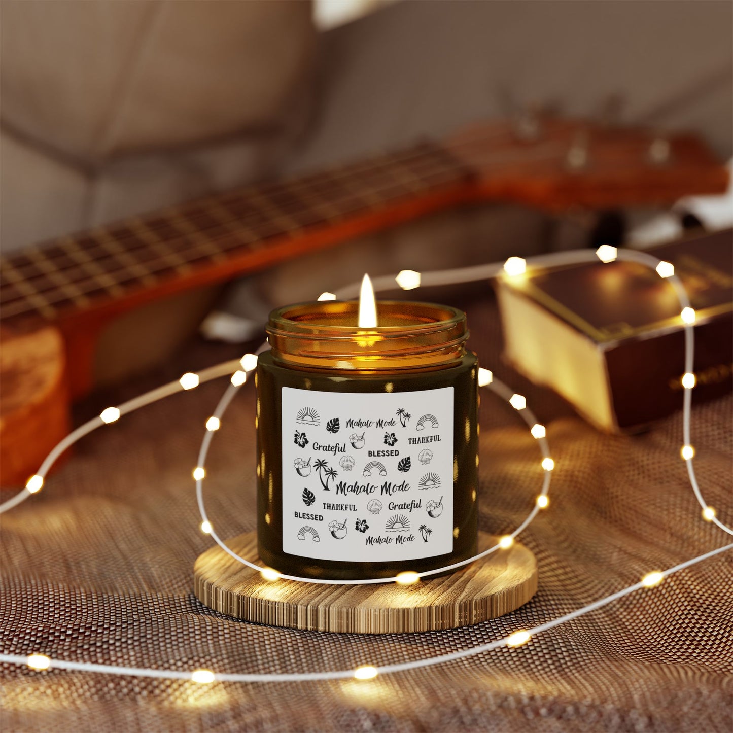 A dark amber glass jar candle with a gold metal screw-on lid and a white paper label featuring black hand-drawn style illustrations and the words 'Mahalo • Mode', 'Grateful', 'BLESSED', and 'THANKFUL'.on a wooden coaster, surrounded by string lights and a guitar in the background.