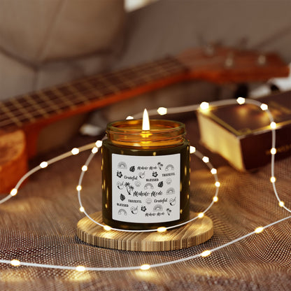 A dark amber glass jar candle with a gold metal screw-on lid and a white paper label featuring black hand-drawn style illustrations and the words 'Mahalo • Mode', 'Grateful', 'BLESSED', and 'THANKFUL'.on a wooden coaster, surrounded by string lights and a guitar in the background.