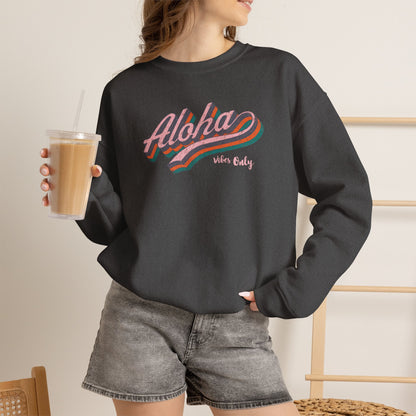 A woman wearing a dark charcoal gray crewneck sweatshirt with a colorful retro-style 'Aloha vibes only' graphic, paired with gray denim shorts and holding an iced coffee.