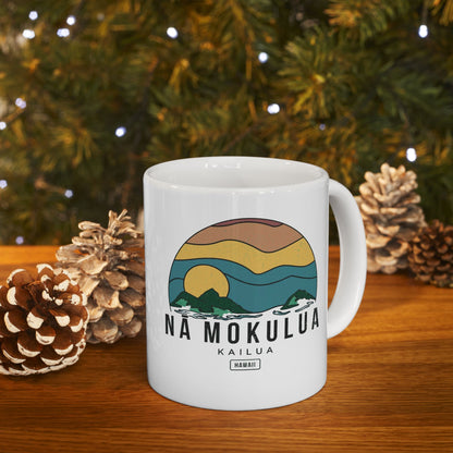 A white ceramic mug with a colorful retro sunset landscape graphic and the text 'NA MOKULUA KAILUA HAWAII' on the front, displayed on a wooden surface with pinecones and blurred Christmas tree lights in the background.