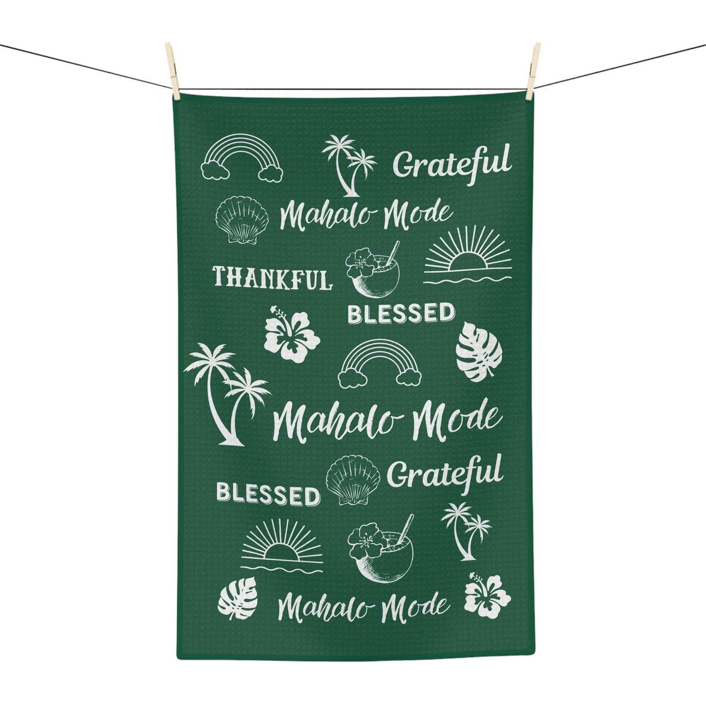A dark green rectangular kitchen towel or tea towel hanging from a clothesline with wooden clips, featuring white tropical-themed illustrations and phrases including 'Mahalo Mode', 'Blessed', 'Grateful', palm trees, hibiscus flowers, monstera leaves, rainbows, shells, and sunsets.