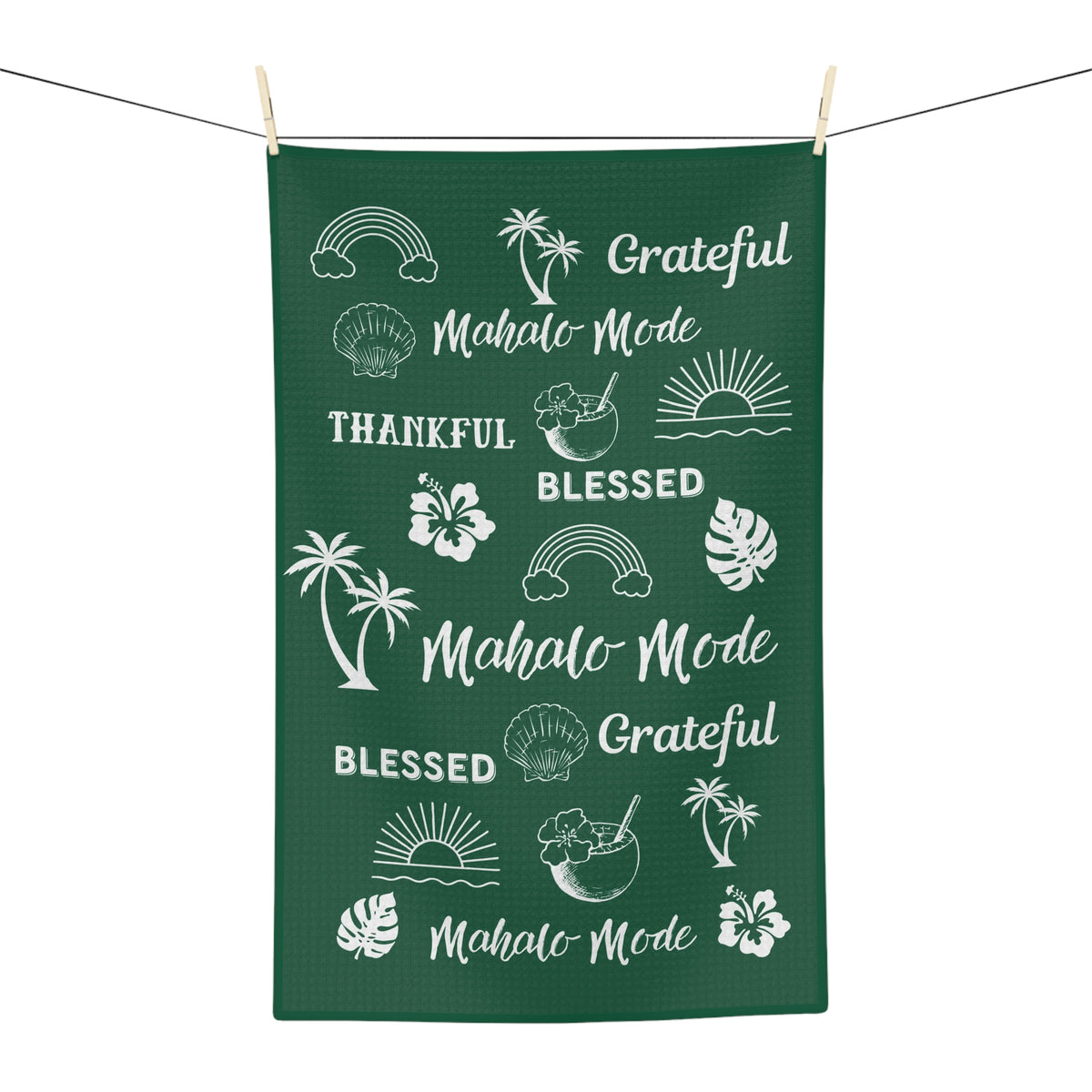 A dark green rectangular kitchen towel or tea towel hanging from a clothesline with wooden clips, featuring white tropical-themed illustrations and phrases including 'Mahalo Mode', 'Blessed', 'Grateful', palm trees, hibiscus flowers, monstera leaves, rainbows, shells, and sunsets.