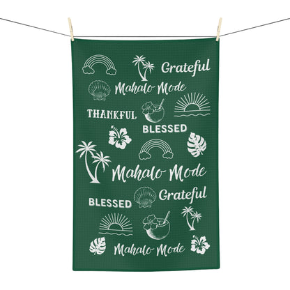 A dark green rectangular kitchen towel or tea towel hanging from a clothesline with wooden clips, featuring white tropical-themed illustrations and phrases including 'Mahalo Mode', 'Blessed', 'Grateful', palm trees, hibiscus flowers, monstera leaves, rainbows, shells, and sunsets.