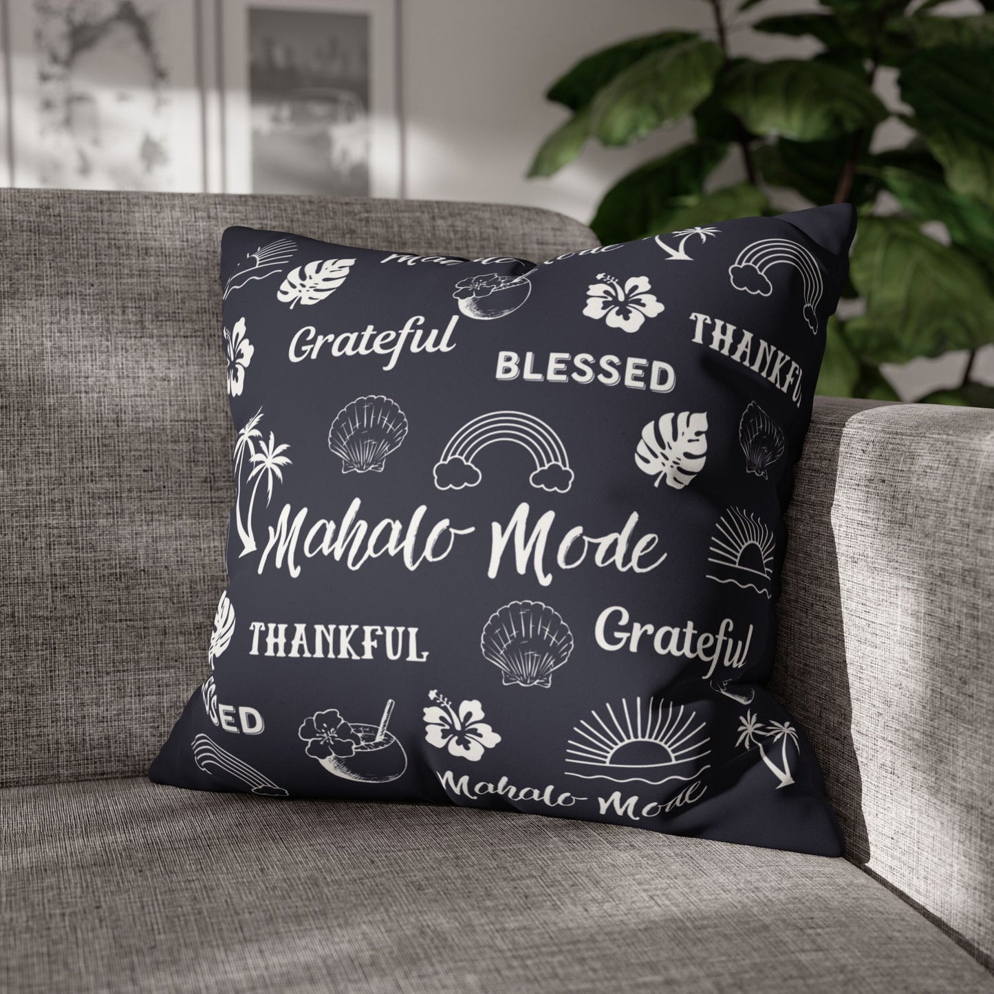 A square throw pillow with a dark navy blue background and white tropical-themed illustrations and text, resting on a gray fabric sofa in a living room setting with a plant in the background.