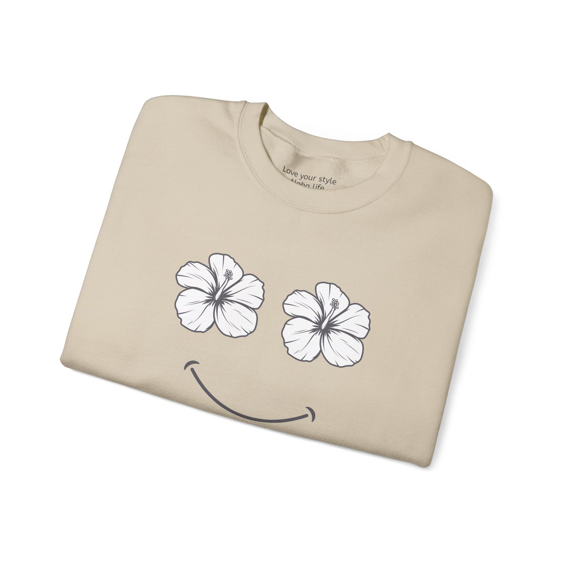 A beige crewneck t-shirt folded neatly on a white background, featuring a graphic of two white hibiscus flowers forming eyes and a black smiley mouth.