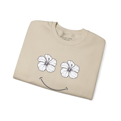 A beige crewneck t-shirt folded neatly on a white background, featuring a graphic of two white hibiscus flowers forming eyes and a black smiley mouth.