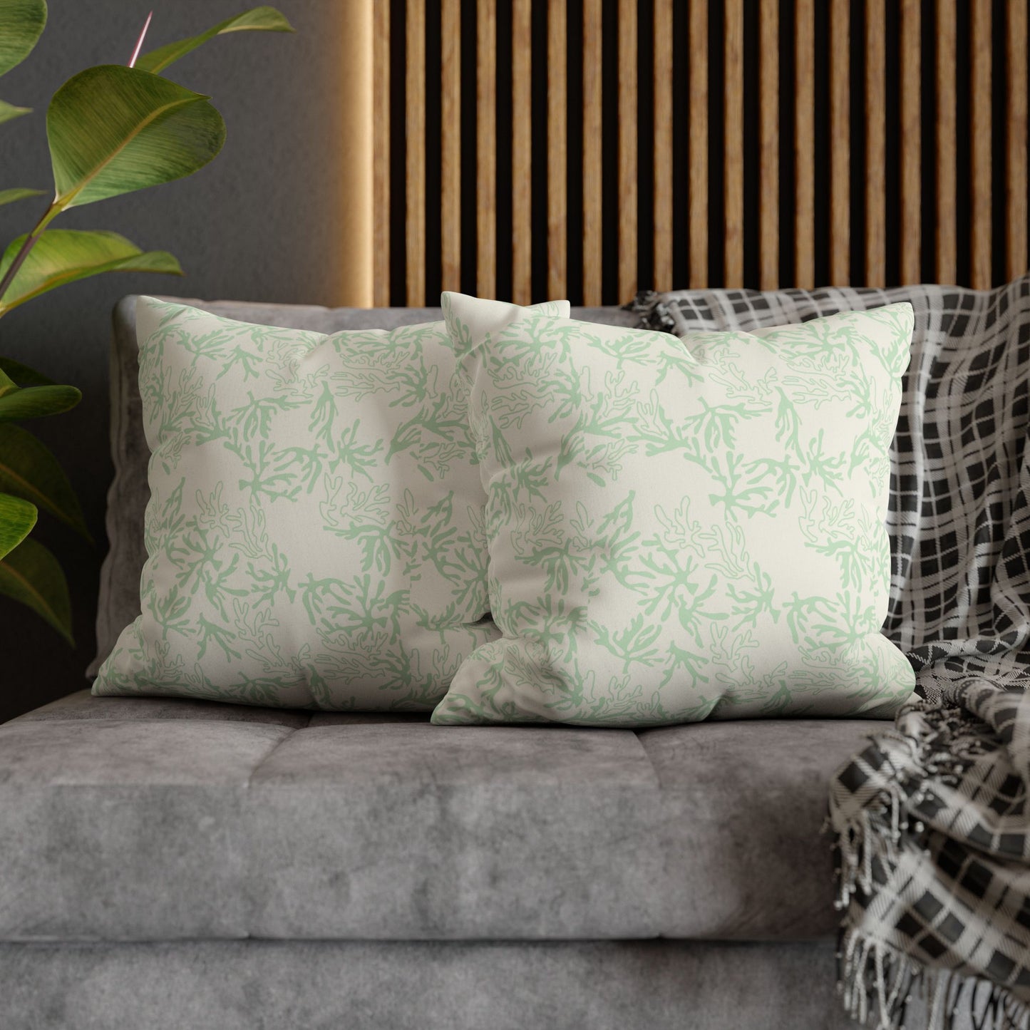 Two square throw pillow with a light sage green coral pattern on an off-white background, resting on a textured gray fabric sofa with a green houseplant in the background.