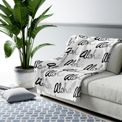 Aloha grayscale sherpa fleece blanket with a repeating black, white and gray island-inspired design draped over a grey couch.