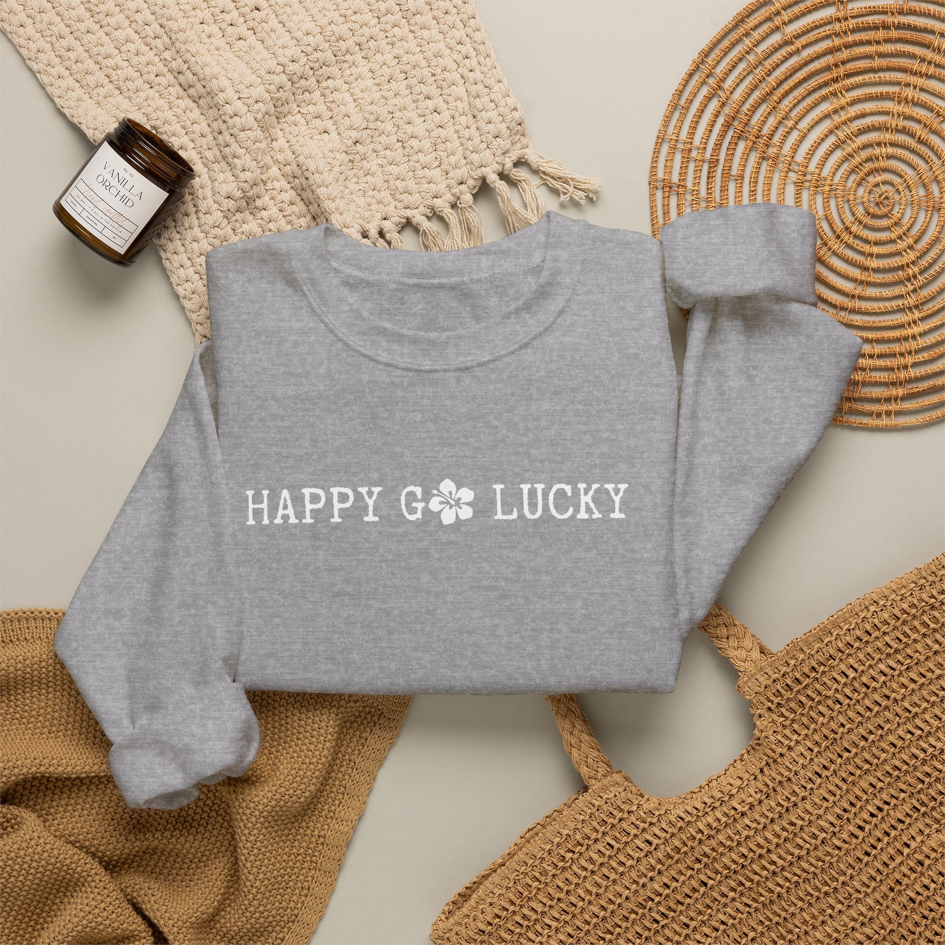 Gray sweatshirt with 'HAPPY GO LUCKY text on a textured surface with a bottle and woven mat.
