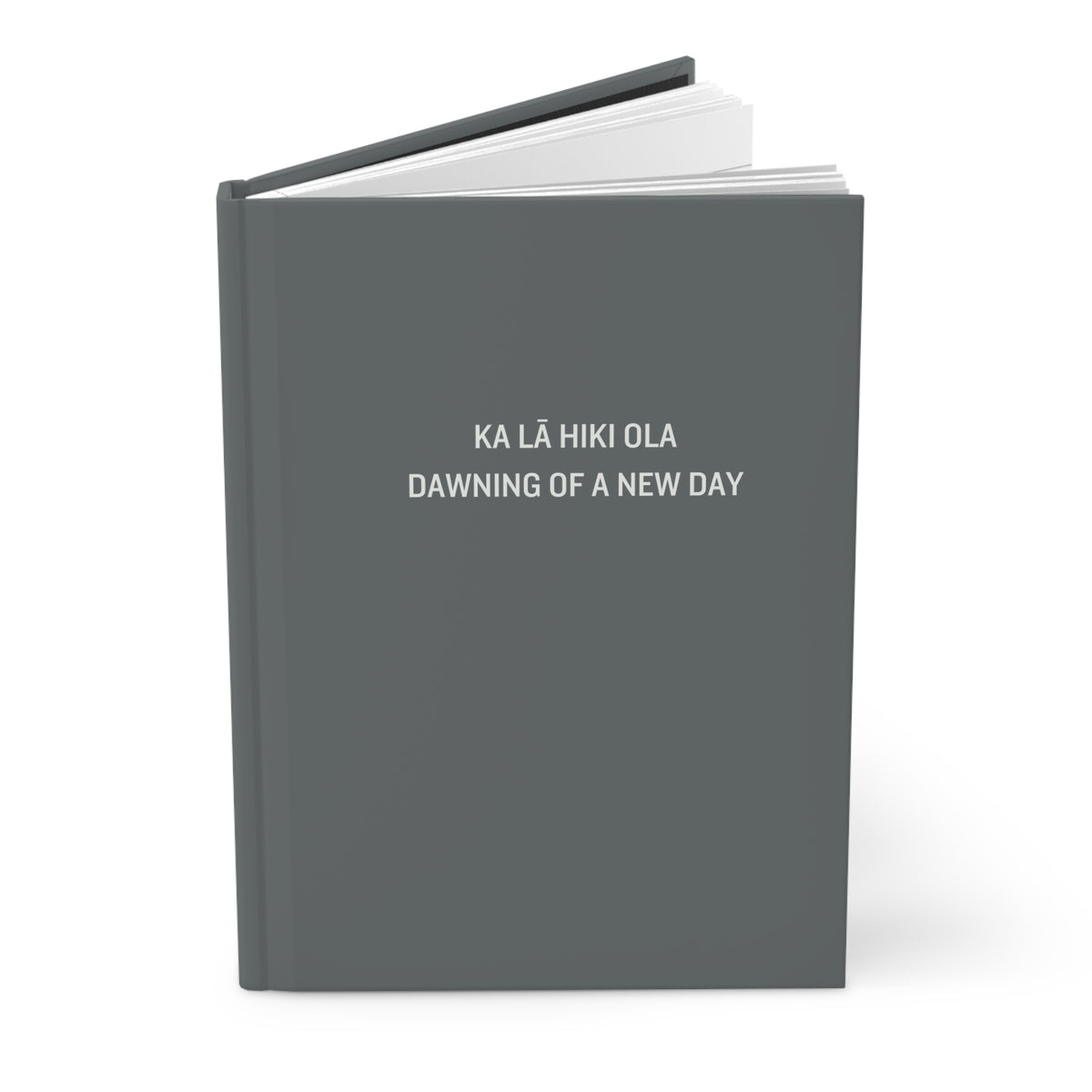 A hardcover notebook or journal shown closed at a slight angle, with a dark gray cover and white text reading 'KA LÁ HIKI OLA DAWNING OF A NEW DAY' on a clean white background.