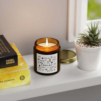 A dark amber glass jar candle with a gold metal screw-on lid and a white paper label featuring black hand-drawn style illustrations and the words 'Mahalo • Mode', 'Grateful', 'BLESSED', and 'THANKFUL' sitting on a white table with a plant and books.