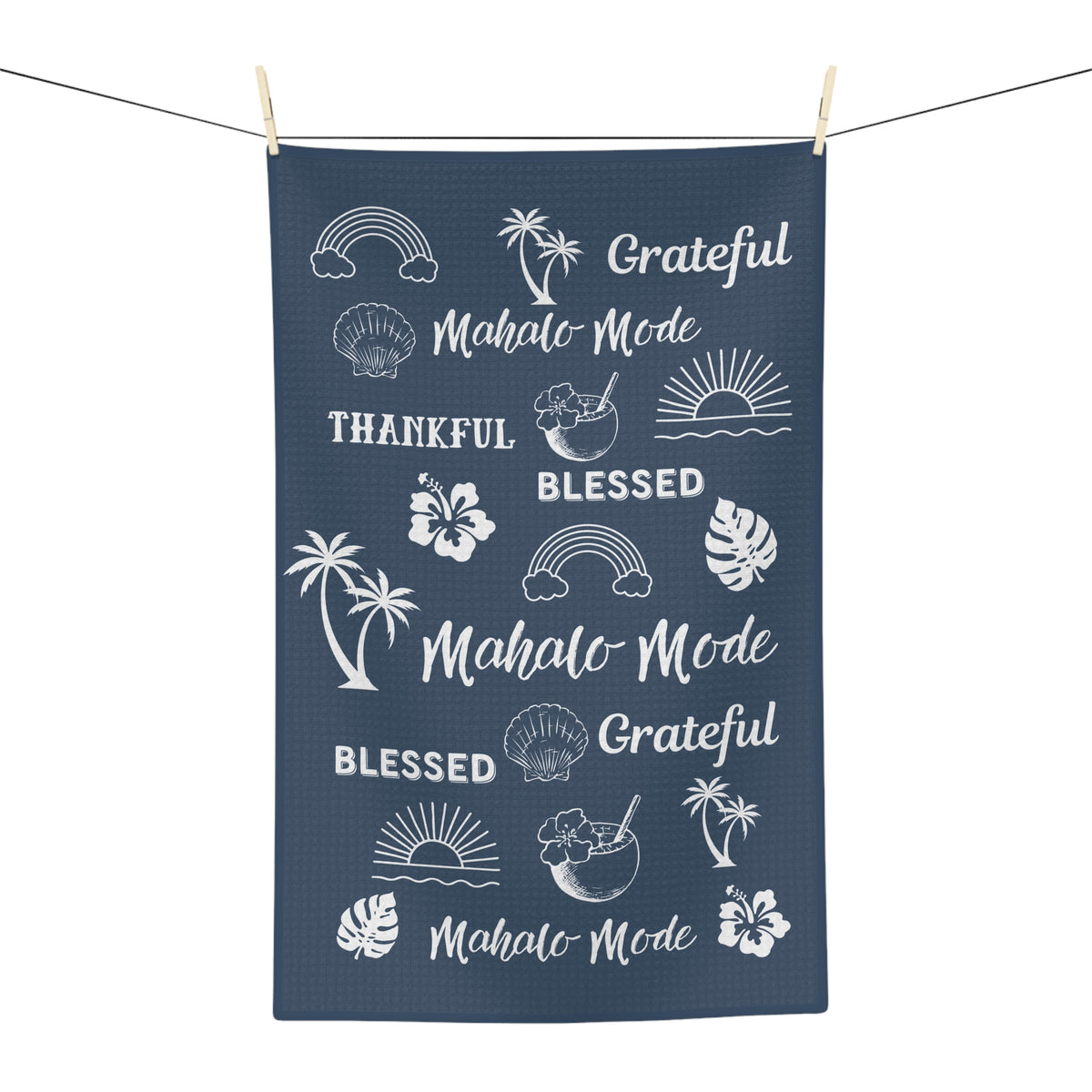 A navy blue kitchen towel with white tropical-themed graphics and text including 'Mahalo Mode', 'Blessed', and 'Grateful', hanging from a string with wooden clothespins against a white background.