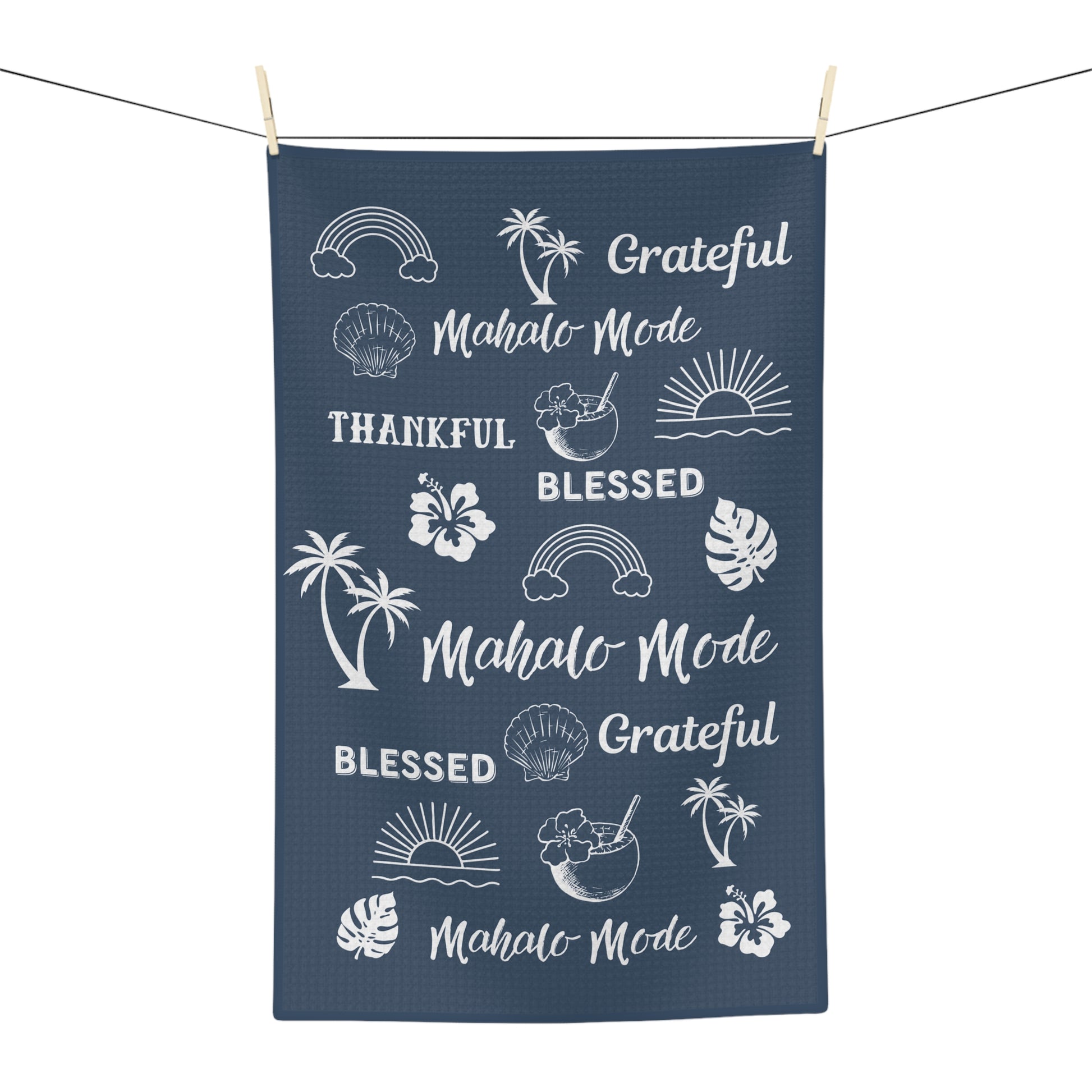 A navy blue kitchen towel with white tropical-themed graphics and text including 'Mahalo Mode', 'Blessed', and 'Grateful', hanging from a string with wooden clothespins against a white background.