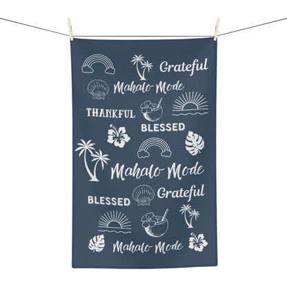 A navy blue kitchen towel with white tropical-themed graphics and text including 'Mahalo Mode', 'Blessed', and 'Grateful', hanging from a string with wooden clothespins against a white background.