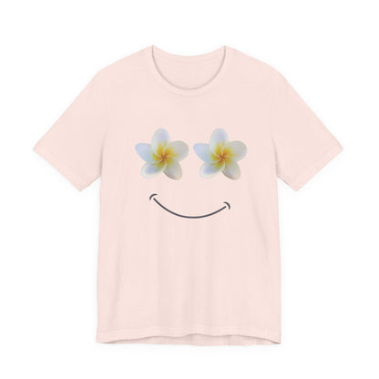 A light pink short-sleeve crewneck t-shirt featuring a graphic of two white plumeria flowers with yellow centers as eyes and a simple black smiley curve below, displayed on a clean white background.