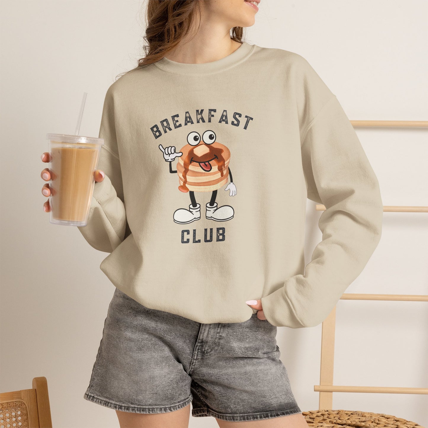 A woman wearing a beige crewneck sweatshirt with a cartoon pancake graphic and the text 'BREAKFAST CLUB'. She is holding an iced coffee and wearing gray denim shorts in a brightly lit indoor setting.