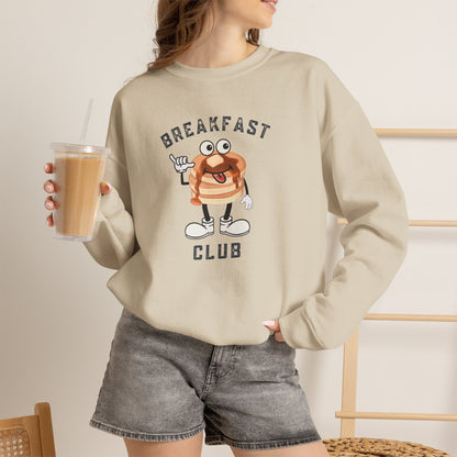 A woman wearing a beige crewneck sweatshirt with a cartoon pancake graphic and the text 'BREAKFAST CLUB'. She is holding an iced coffee and wearing gray denim shorts in a brightly lit indoor setting.