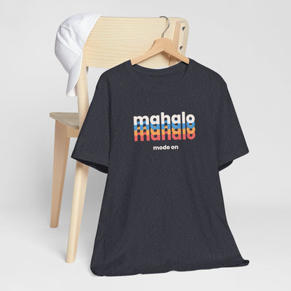 A dark navy heather short-sleeve t-shirt on a wooden hanger, styled in front of a light wood chair with a white cap draped over the backrest. The shirt features a retro-style graphic on the chest reading 'mahalo mode' in white and rainbow stripes, with 'mode on' printed below.