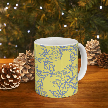 A white ceramic mug with a yellow and blue coral pattern, set on a wooden table with pinecones and a blurred Christmas tree background.