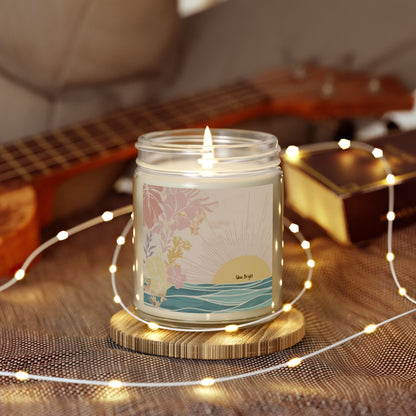 A lit glass jar candle with a tropical sunset and floral label sits on a wooden coaster, surrounded by warm fairy lights, with a guitar and books blurred in the background.