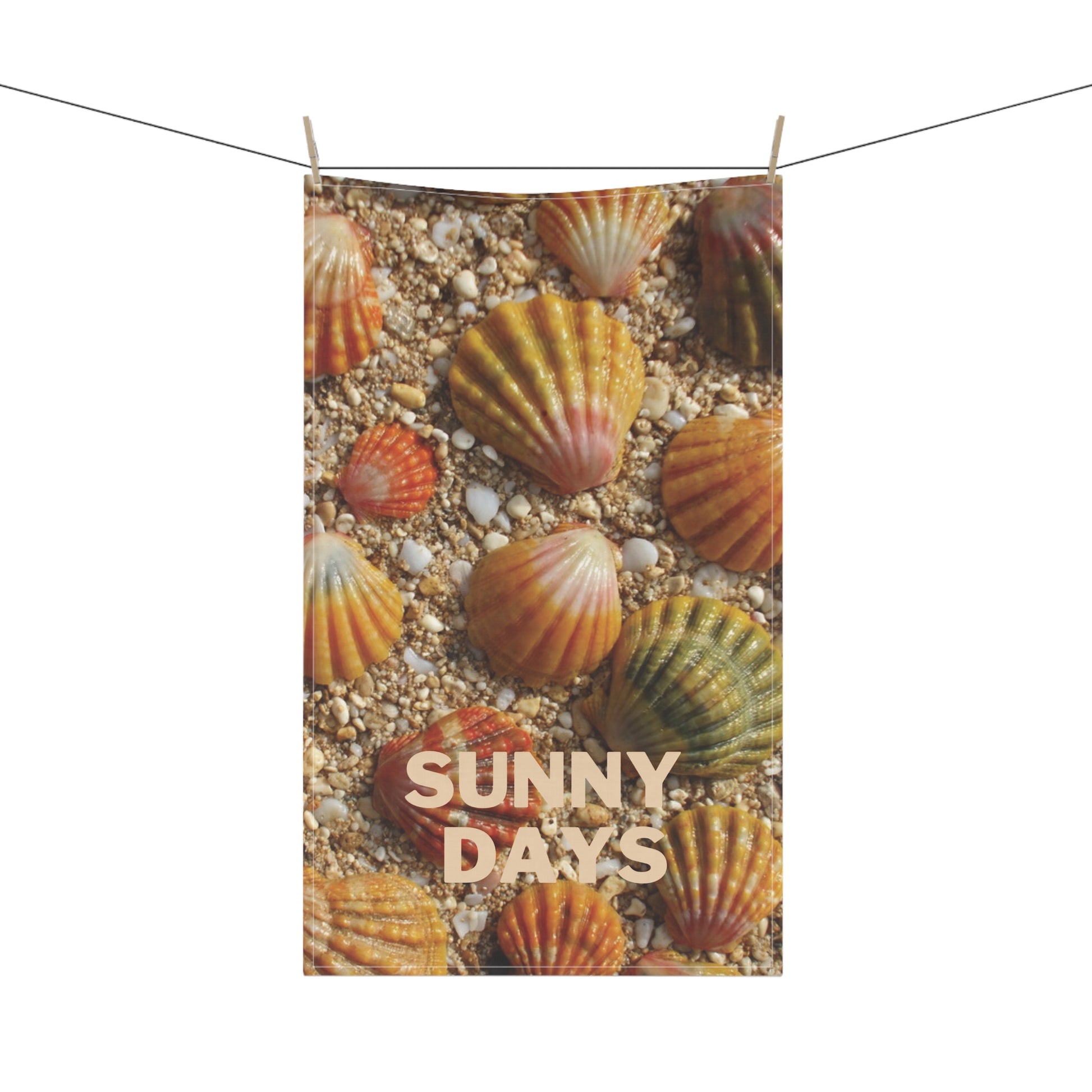 A rectangular teatowel printed with seashells on sandy ground and the words “SUNNY DAYS,” hanging from a string with two wooden clips against a white background.