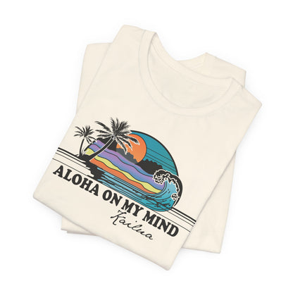 A folded cream-colored t-shirt with a retro-style graphic of a sunset over palm trees and waves, with the text 'ALOHA ON MY MIND' and 'Kailua' printed below.