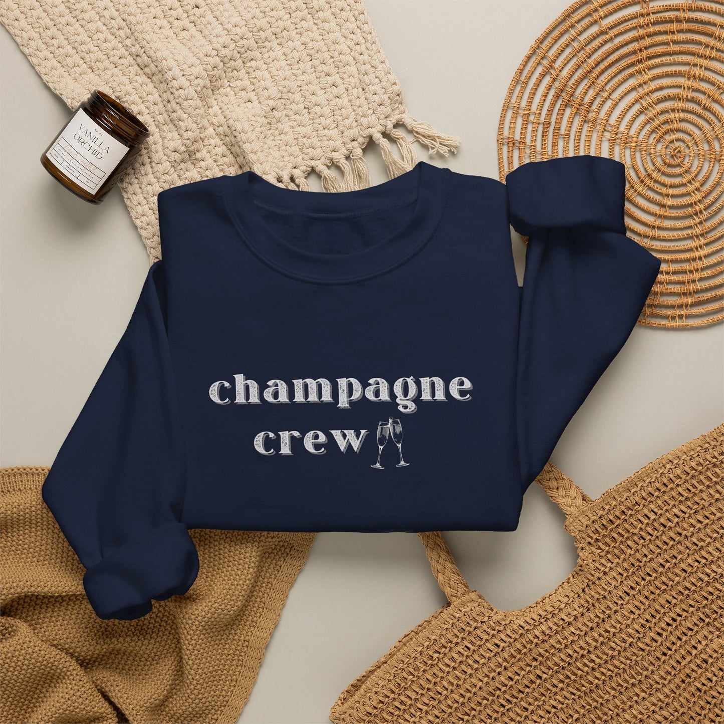 A navy crewneck sweatshirt with the word 'champagne crew' printed in white lowercase serif letters across the chest, styled in a flat lay with a woven cream throw, a small amber candle jar, and a woven mat on a light beige background.