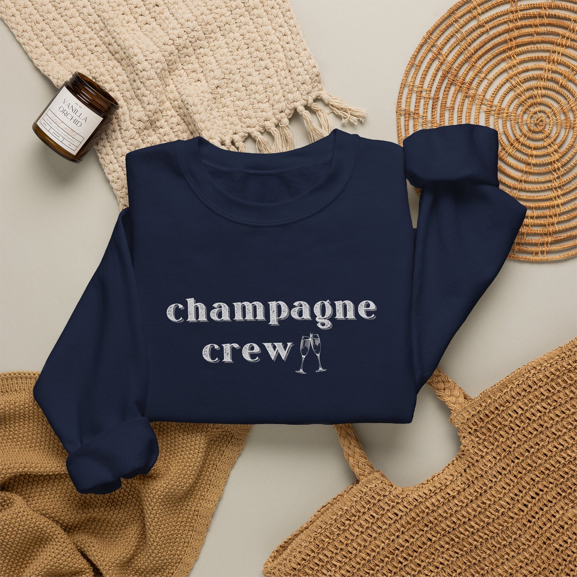 A navy crewneck sweatshirt with the word 'champagne crew' printed in white lowercase serif letters across the chest, styled in a flat lay with a woven cream throw, a small amber candle jar, and a woven mat on a light beige background.