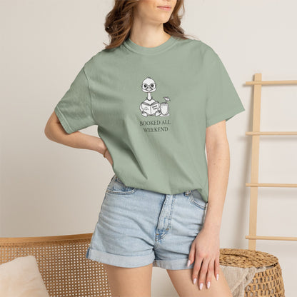 A woman standing wearing a light green comfort colors t-shirt with design of a nene goose reading a book and text 'booked all weekend' paired with denim shorts
