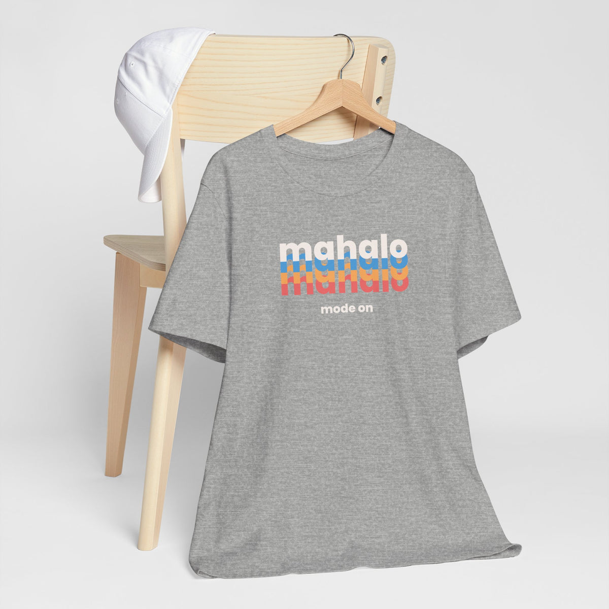 An athletic heather short-sleeve t-shirt on a wooden hanger, styled in front of a light wood chair with a white cap draped over the backrest. The shirt features a retro-style graphic on the chest reading 'mahalo mode' in white and rainbow stripes, with 'mode on' printed below.
