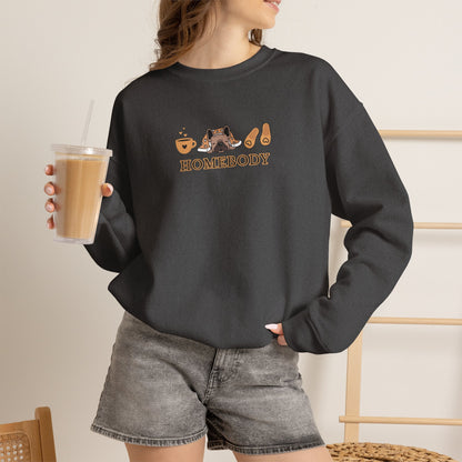 Person holding an iced coffee standing wearing a dark grey crewneck sweatshirt featuring a central graphic of a dog under a blanket, house slippers, coffee cups and the word 'HOMEBODY' printed below it.