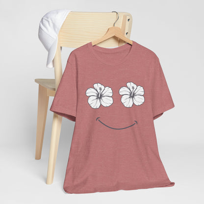 A heathered dusty rose t-shirt on a wooden hanger, featuring a graphic of two white hibiscus flowers forming eyes and a simple smile. The shirt is styled leaning against a light wood chair with a white t-shirt draped over the back, set against a clean light gray background.