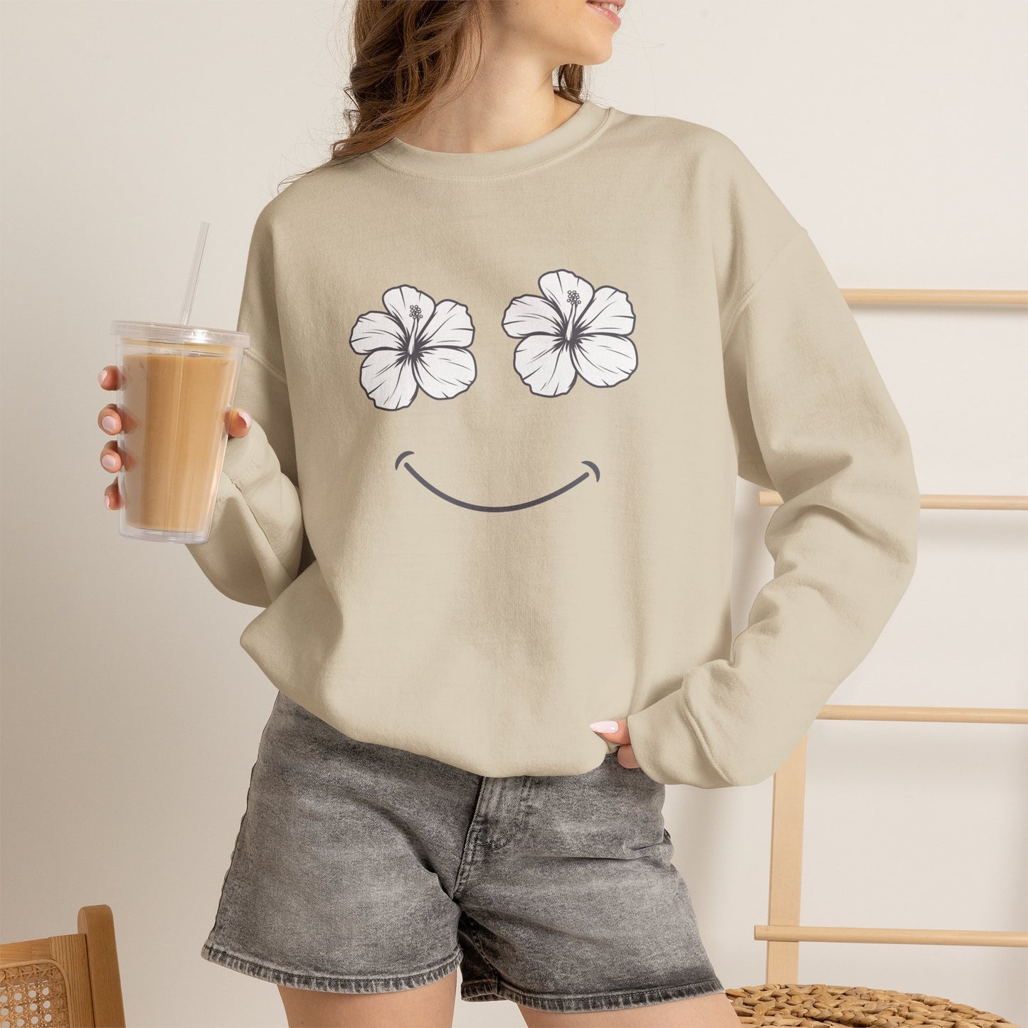 Person wearing a Sand Happy Hibiscus Sweatshirt featuring a cheerful hibiscus flower smile design inspired by island life and aloha style holding a drink.