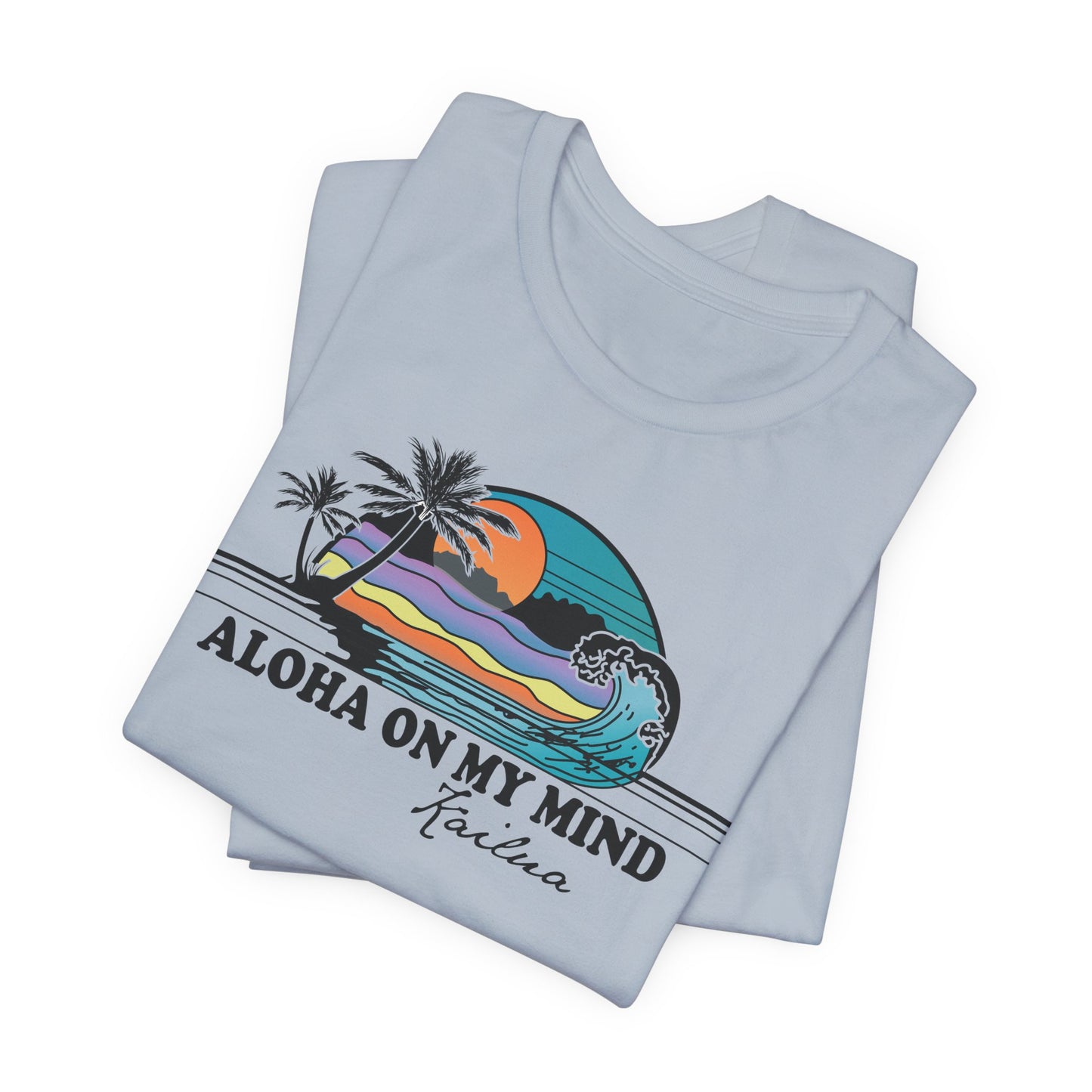 A folded light blue t-shirt with a colorful retro graphic of a Hawaiian sunset and wave, featuring the text 'ALOHA ON MY MIND' and 'Kailua'.