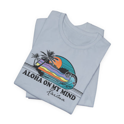 A folded light blue t-shirt with a colorful retro graphic of a Hawaiian sunset and wave, featuring the text 'ALOHA ON MY MIND' and 'Kailua'.