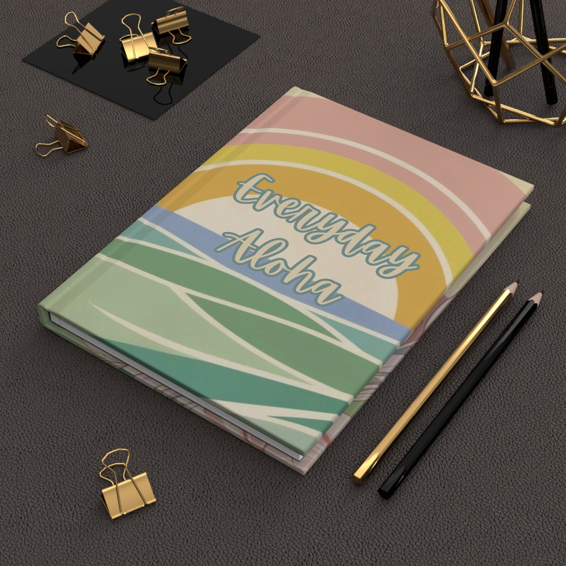 A notebook with a pastel rainbow cover design reading 'Everyday Aloha' lies on a dark textured surface, styled with gold binder clips, pencils, and a geometric gold decor object.