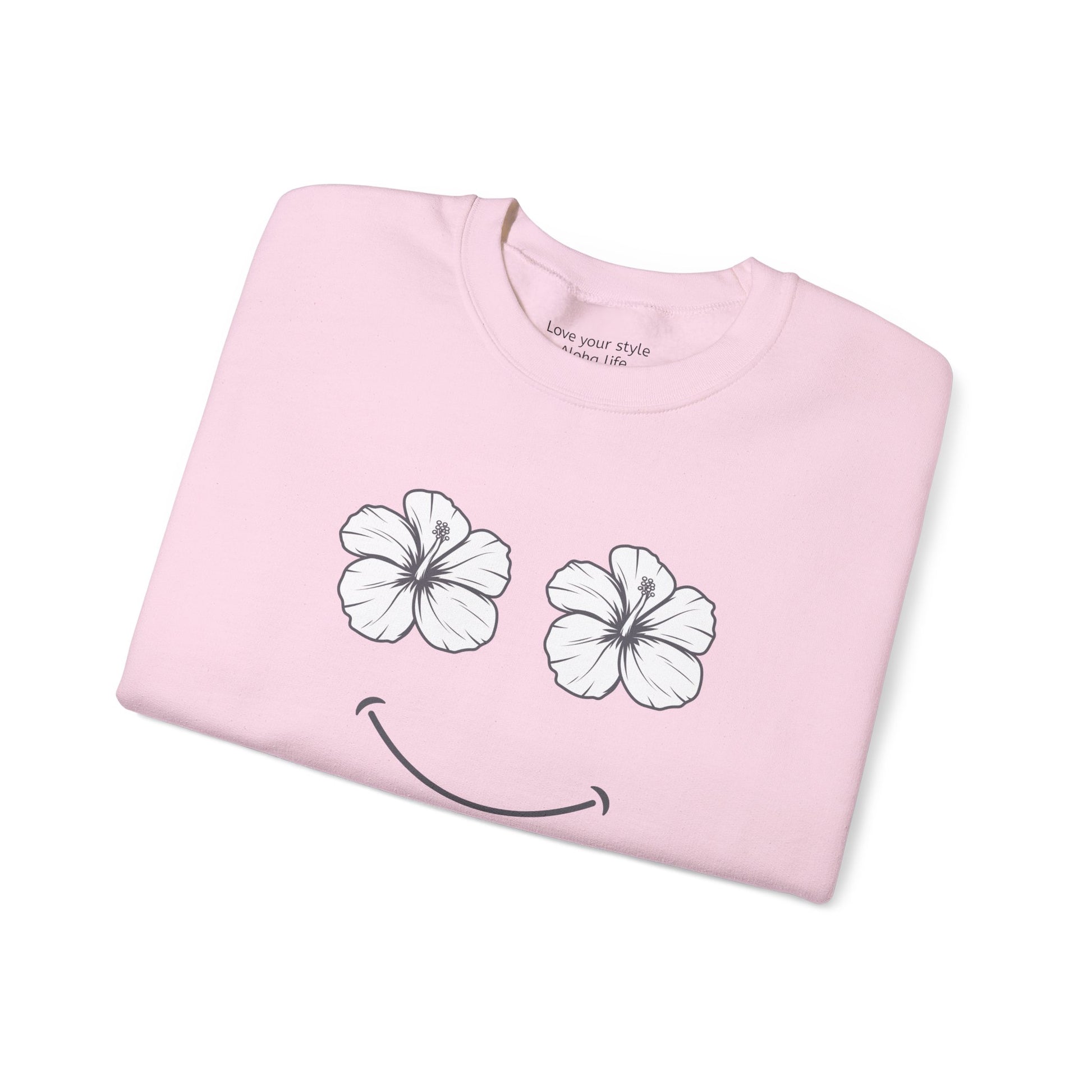 A folded light pink t-shirt with a graphic of two white hibiscus flowers forming eyes and a black smiley mouth, shown on a plain white background.