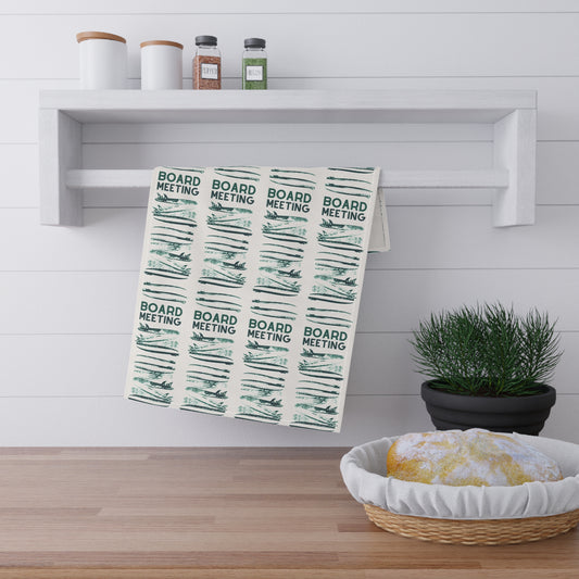 A kitchen towel with a repeating green and black 'BOARD MEETING' pattern hanging from a white rack on a white shiplap wall, with a wooden floor, spice jars, a potted plant, and a basket of bread in the scene.