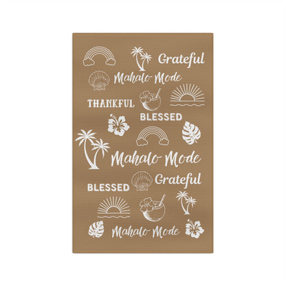 A tan kitchen towel with white tropical-themed illustrations and phrases including rainbows, palm trees, hibiscus flowers, seashells, and repeated text reading 'Grateful Mahalo Mode' and 'Blessed.'.