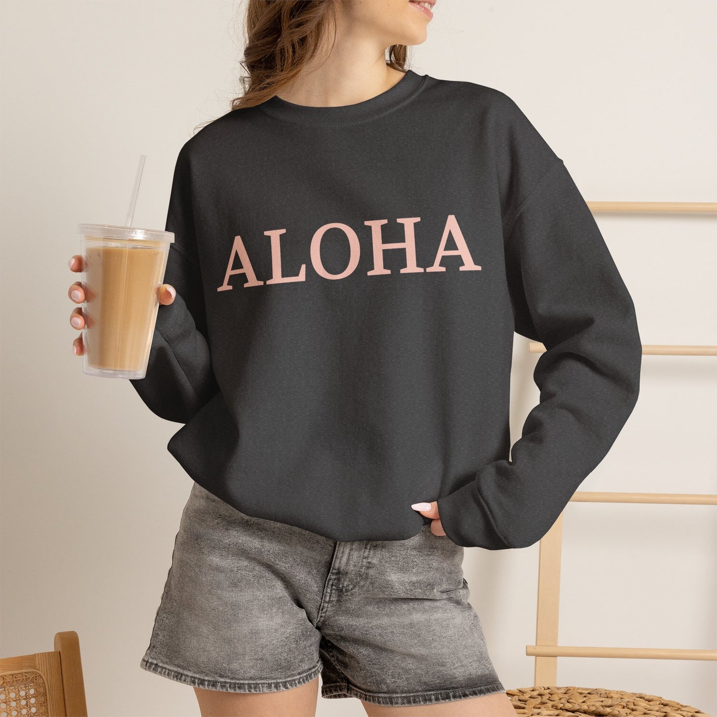 A woman wearing an oversized crewneck sweatshirt in dark grey with the word 'ALOHA' printed across the chest in a light pink color. She is holding a clear plastic cup of iced coffee and wearing gray denim shorts, set against a warm, neutral indoor background.