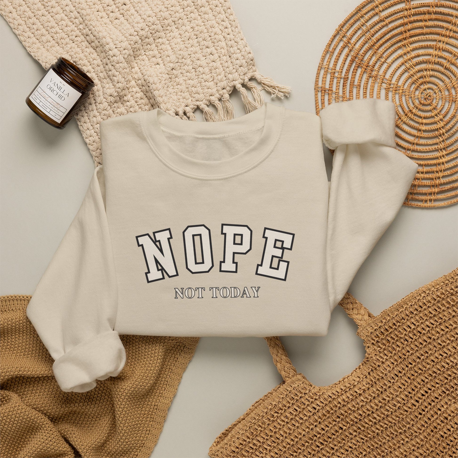 A sand crewneck sweatshirt with the words 'NOPE NOT TODAY' printed on the front, styled in a flat lay with a beige knit blanket, a woven basket, and a small candle.