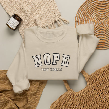 A sand crewneck sweatshirt with the words 'NOPE NOT TODAY' printed on the front, styled in a flat lay with a beige knit blanket, a woven basket, and a small candle.